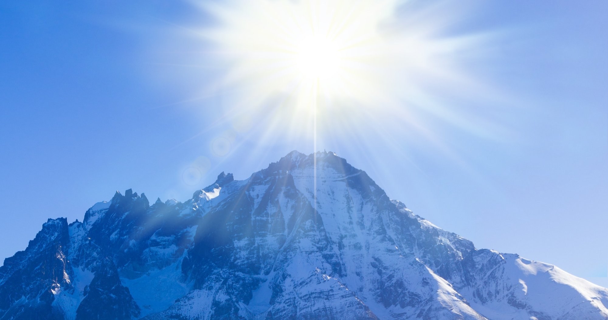 Sunlight bursting directly over the summit of a snow covered mountain range against a clear blue sky.